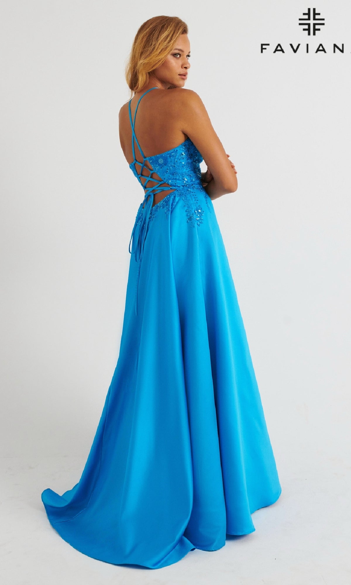 A woman in the Faviana 11260—a vibrant blue, floor-length satin prom dress with a lace-embroidered bodice and lace-up back—stands looking over her shoulder. The FAVIAN logo is displayed in the top right corner.